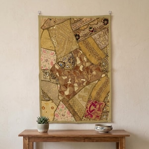 May include: A rectangular wall hanging featuring a patchwork of various fabrics in shades of gold, beige, and brown. The textile includes intricate embroidery, sequins, metallic thread, and small floral patterns, finished with a solid gold border.