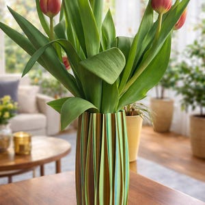 Könnte beinhalten: Eine dekorative Vase mit vertikalem Streifendesign in Grün-, Braun- und Blautönen hält einen Strauß Tulpen. Die Tulpen haben rote und gelbe Blütenblätter und lange grüne Blätter. Die Vase steht auf einem Holztisch.