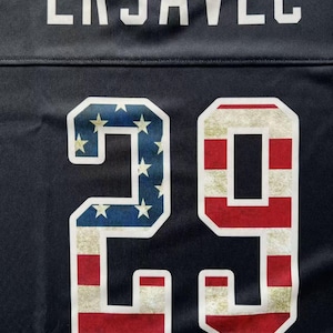 K&ouml;nnte beinhalten: Nahaufnahme eines marineblauen Trikots mit dem Namen "ERJAVEC" in wei&szlig;en Buchstaben. Die Nummer "29" ist mit einem patriotischen Design mit der amerikanischen Flagge versehen.