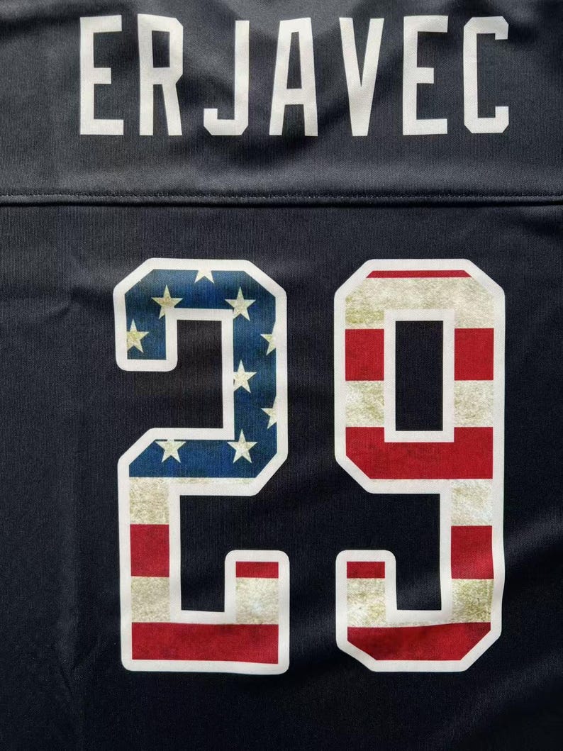 K&ouml;nnte beinhalten: Nahaufnahme eines marineblauen Trikots mit dem Namen "ERJAVEC" in wei&szlig;en Buchstaben. Die Nummer "29" ist mit einem patriotischen Design mit der amerikanischen Flagge versehen.