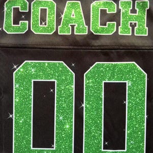 May include: A black jersey featuring the word COACH in large, green glittery block letters across the top. Below the text, the number 00 is displayed in the same green glittery material with a white outline, set against the black fabric.