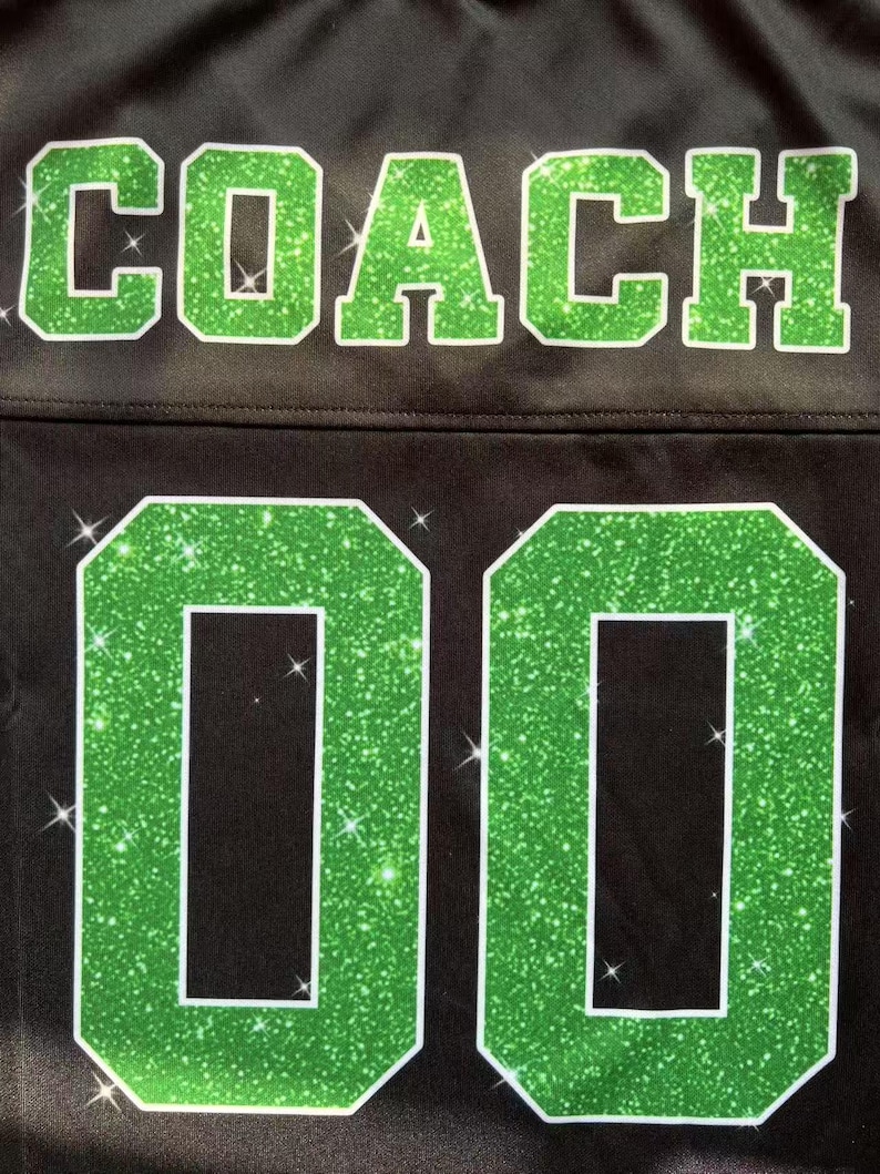 May include: A black jersey featuring the word COACH in large, green glittery block letters across the top. Below the text, the number 00 is displayed in the same green glittery material with a white outline, set against the black fabric.
