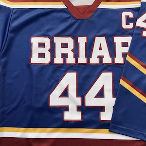 K&ouml;nnte beinhalten: Ein blaues Hockey-Trikot mit dem Wort "BRIAF" und der Nummer "44" in wei&szlig;en Blockbuchstaben. Das Trikot hat einen weinroten und wei&szlig;en Saum, mit goldenen und wei&szlig;en Streifen auf den &Auml;rmeln und Schultern. Der Buchstabe "C" ist sichtbar.