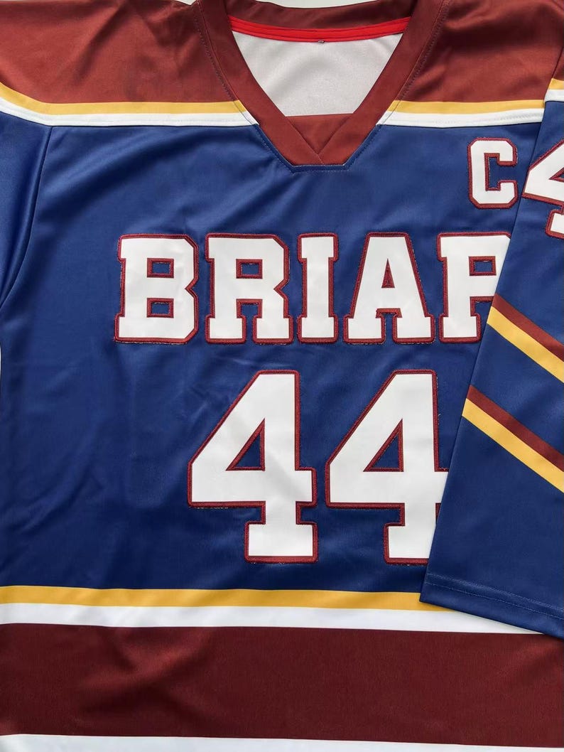 K&ouml;nnte beinhalten: Ein blaues Hockey-Trikot mit dem Wort "BRIAF" und der Nummer "44" in wei&szlig;en Blockbuchstaben. Das Trikot hat einen weinroten und wei&szlig;en Saum, mit goldenen und wei&szlig;en Streifen auf den &Auml;rmeln und Schultern. Der Buchstabe "C" ist sichtbar.