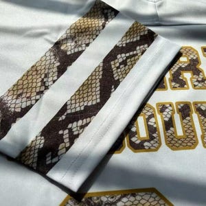 May include: A white athletic-style shirt featuring a sleeve with two horizontal stripes in a brown and tan snakeskin pattern. The fabric displays partial text with block lettering filled with the same snakeskin print and outlined in gold.