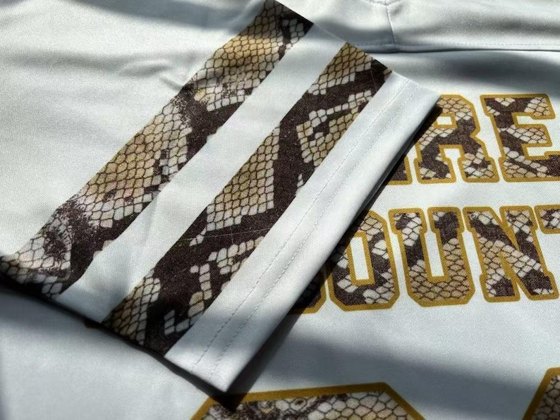 May include: A white athletic-style shirt featuring a sleeve with two horizontal stripes in a brown and tan snakeskin pattern. The fabric displays partial text with block lettering filled with the same snakeskin print and outlined in gold.