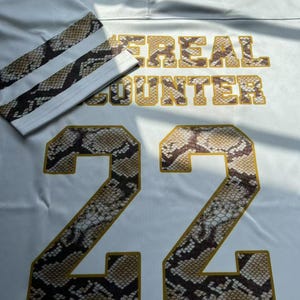 May include: White football jersey with the number 22 in a snakeskin pattern. The jersey features the text "#REAL COUNTER" in gold lettering. The sleeves have a snakeskin pattern with white stripes.