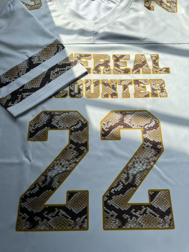 May include: White football jersey with the number 22 in a snakeskin pattern. The jersey features the text "#REAL COUNTER" in gold lettering. The sleeves have a snakeskin pattern with white stripes.