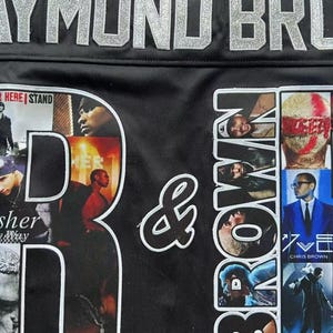 May include: A black jersey featuring large silver glitter lettering that reads RAYMOND BROWN. The letter R contains photo collages of Usher, while the letter B contains photo collages of Chris Brown. A white ampersand sits between the letters.