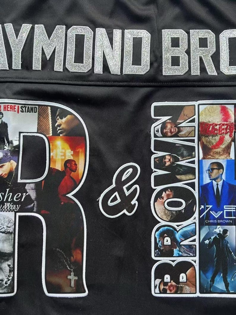 May include: A black jersey featuring large silver glitter lettering that reads RAYMOND BROWN. The letter R contains photo collages of Usher, while the letter B contains photo collages of Chris Brown. A white ampersand sits between the letters.