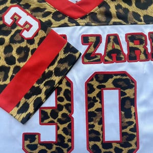 May include: A white sports jersey featuring a leopard print pattern on the shoulders and sleeves. The jersey has a red V-neck collar, red sleeve stripes, and the number 30 in leopard print with red outlines. The text ZAR is visible on the front.