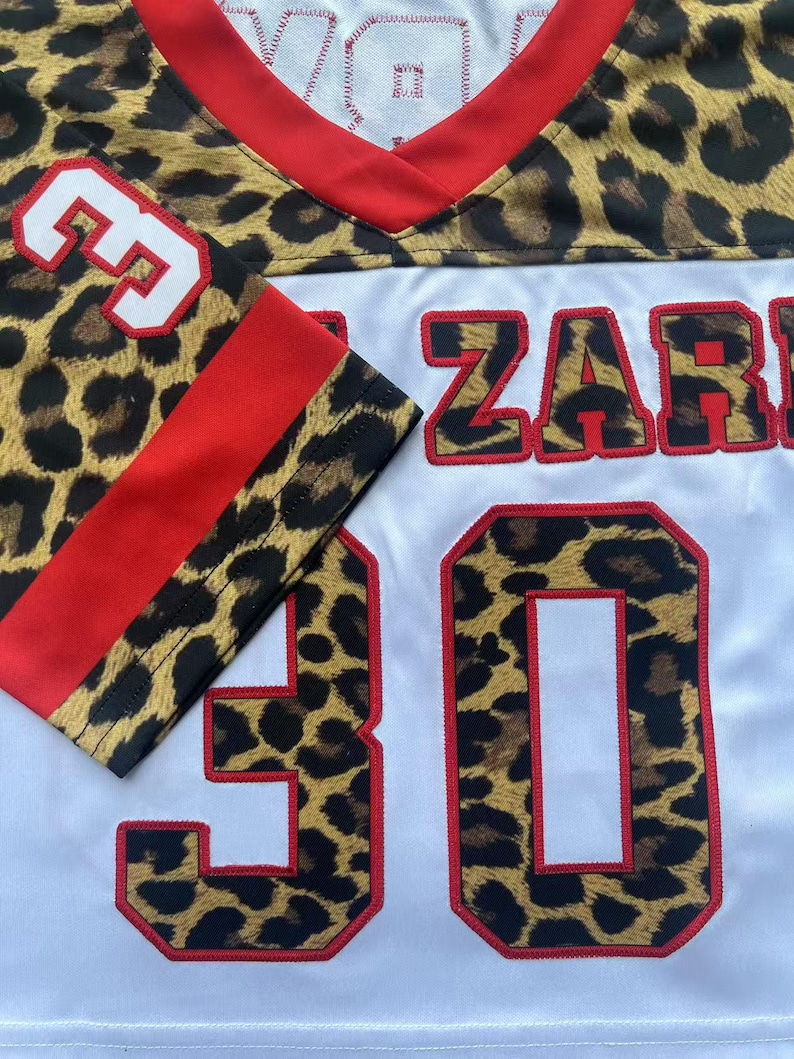 May include: A white sports jersey featuring a leopard print pattern on the shoulders and sleeves. The jersey has a red V-neck collar, red sleeve stripes, and the number 30 in leopard print with red outlines. The text ZAR is visible on the front.