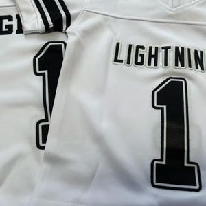 Pu&ograve; includere: Due maglie sportive bianche con scritte nere che riportano LIGHTNINGS e il numero 1 in un carattere grassetto. Le maglie presentano uno scollo a V e strisce nere sulle maniche, con il testo e i numeri applicati in un chiaro stile a blocchi.