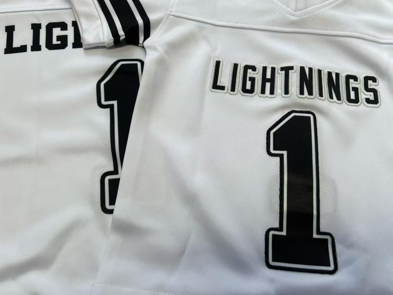Pu&ograve; includere: Due maglie sportive bianche con scritte nere che riportano LIGHTNINGS e il numero 1 in un carattere grassetto. Le maglie presentano uno scollo a V e strisce nere sulle maniche, con il testo e i numeri applicati in un chiaro stile a blocchi.