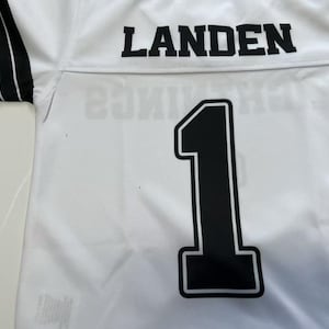 Pu&ograve; includere: Una maglia sportiva bianca con il nome LANDEN in lettere stampatello nere sulla parte superiore della schiena. Sotto il nome, un grande numero 1 nero con un bordo bianco &egrave; centrato. Le maniche corte presentano strisce nere con righe bianche.