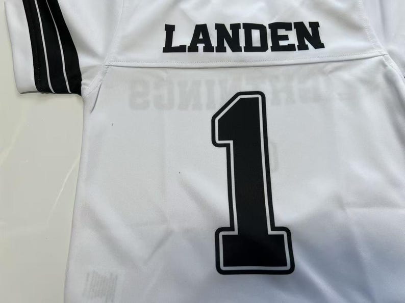 Pu&ograve; includere: Una maglia sportiva bianca con il nome LANDEN in lettere stampatello nere sulla parte superiore della schiena. Sotto il nome, un grande numero 1 nero con un bordo bianco &egrave; centrato. Le maniche corte presentano strisce nere con righe bianche.