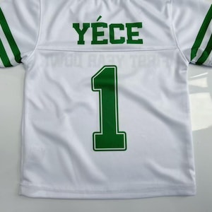 Pu&ograve; includere: Una maglia bianca a maniche corte con strisce verdi sulle maniche, il nome Y&Eacute;CE stampato in verde sulla parte superiore della schiena e il numero 1 in un grande carattere verde centrato sotto il nome sul retro del capo.