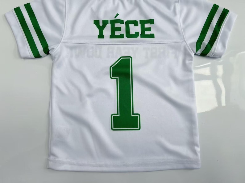 Pu&ograve; includere: Una maglia bianca a maniche corte con strisce verdi sulle maniche, il nome Y&Eacute;CE stampato in verde sulla parte superiore della schiena e il numero 1 in un grande carattere verde centrato sotto il nome sul retro del capo.