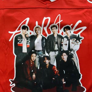 May include: A bright red jersey featuring a large, central graphic patch of seven individuals in dark clothing. The top of the jersey displays white text reading "Tx" with star symbols. The fabric has a V-neck collar with white trim.