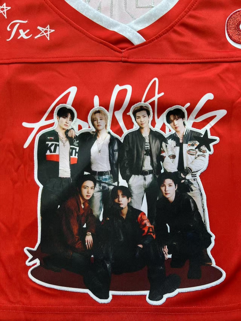 May include: A bright red jersey featuring a large, central graphic patch of seven individuals in dark clothing. The top of the jersey displays white text reading "Tx" with star symbols. The fabric has a V-neck collar with white trim.