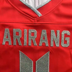 May include: A bright red jersey featuring a V-neck collar with silver trim. The word ARIRANG is printed in large, silver, glittery block letters across the chest, positioned above a silver, glittery, vertical rectangular logo design.