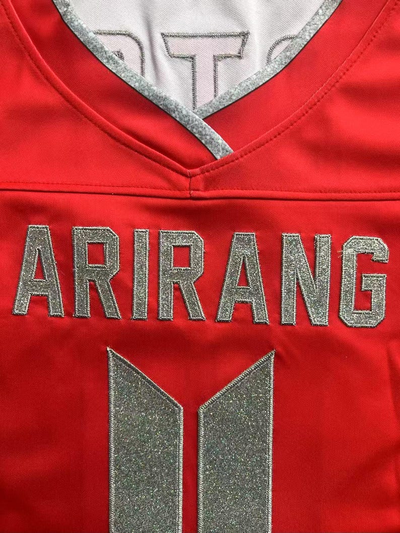 May include: A bright red jersey featuring a V-neck collar with silver trim. The word ARIRANG is printed in large, silver, glittery block letters across the chest, positioned above a silver, glittery, vertical rectangular logo design.