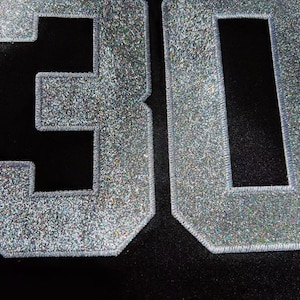 May include: Close-up of the number "30" in silver glitter fabric, stitched onto a black background. The numbers have a textured, sparkling appearance, with a silver outline.