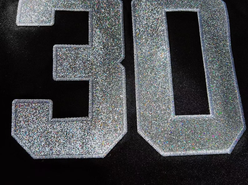 May include: Close-up of the number "30" in silver glitter fabric, stitched onto a black background. The numbers have a textured, sparkling appearance, with a silver outline.