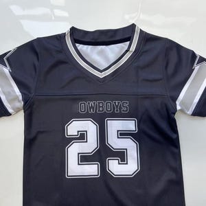Pu&ograve; includere: Una maglia a maniche corte blu navy con scollo a V caratterizzato da strisce bianche e argento. La parte anteriore mostra la scritta OWBOYS sopra il grande numero 25 bianco con un bordo scuro. Ogni manica presenta una stella bianca e argento.
