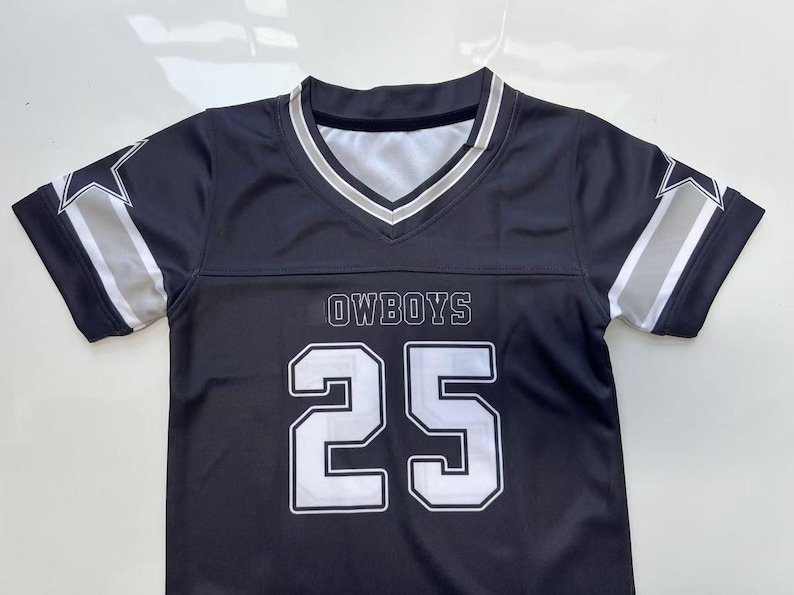 Pu&ograve; includere: Una maglia a maniche corte blu navy con scollo a V caratterizzato da strisce bianche e argento. La parte anteriore mostra la scritta OWBOYS sopra il grande numero 25 bianco con un bordo scuro. Ogni manica presenta una stella bianca e argento.