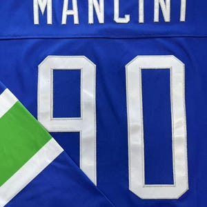 K&ouml;nnte beinhalten: Nahaufnahme eines blauen Trikots mit dem Namen "MANCINI" in wei&szlig;en Buchstaben auf dem oberen R&uuml;cken. Die Nummer "90" ist wei&szlig; auf der Vorderseite zu sehen. Der &Auml;rmel hat gr&uuml;ne und wei&szlig;e Akzente.