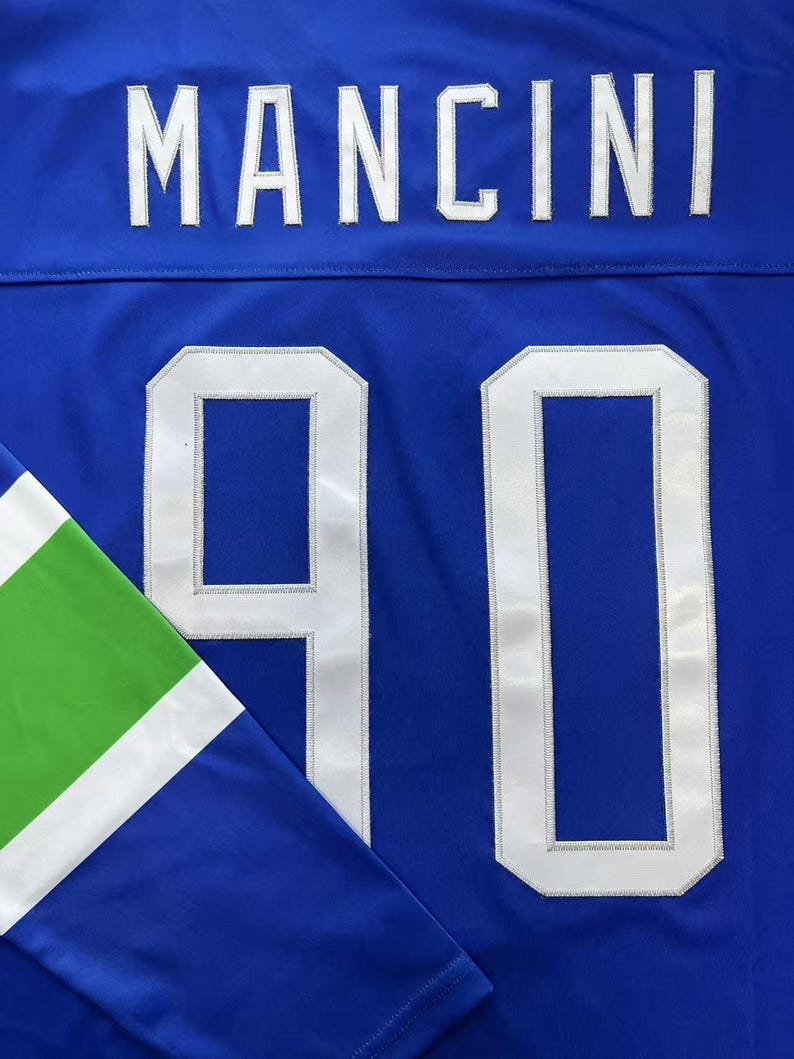 K&ouml;nnte beinhalten: Nahaufnahme eines blauen Trikots mit dem Namen "MANCINI" in wei&szlig;en Buchstaben auf dem oberen R&uuml;cken. Die Nummer "90" ist wei&szlig; auf der Vorderseite zu sehen. Der &Auml;rmel hat gr&uuml;ne und wei&szlig;e Akzente.