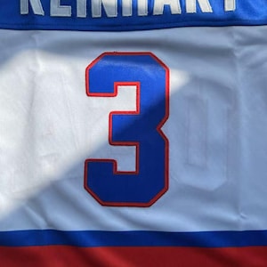 May include: A white, blue, and red hockey jersey with the name "REINHART" in white letters across the top. A large blue number "3" with a red outline is centered on the jersey.
