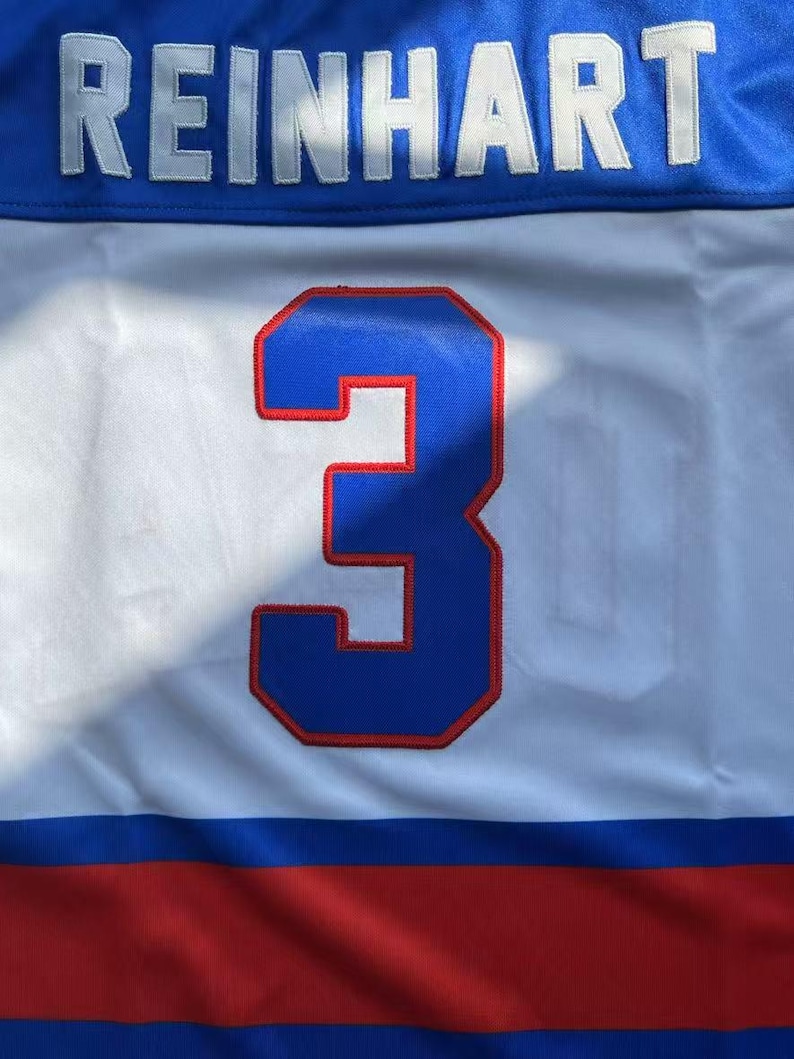 May include: A white, blue, and red hockey jersey with the name "REINHART" in white letters across the top. A large blue number "3" with a red outline is centered on the jersey.