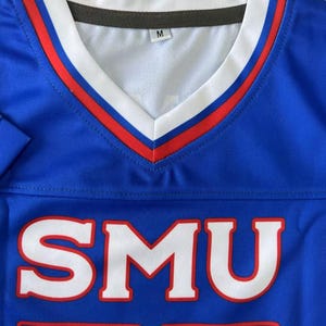 Pu&ograve; includere: Una maglia blu con scollo a V e finiture bianche e rosse. La parte anteriore presenta le lettere SMU in un grande carattere bianco con un contorno rosso. L'etichetta interna sul collo indica la taglia M. Le maniche presentano strisce rosse e bianche.