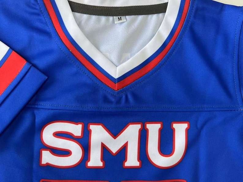 Pu&ograve; includere: Una maglia blu con scollo a V e finiture bianche e rosse. La parte anteriore presenta le lettere SMU in un grande carattere bianco con un contorno rosso. L'etichetta interna sul collo indica la taglia M. Le maniche presentano strisce rosse e bianche.