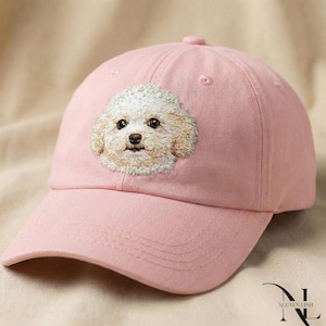May include: A pale pink baseball cap with an embroidered cream-coloured dog's face. The cap has a curved peak and a fabric-covered button. The dog's eyes and nose are dark, and the cap is made of a soft fabric. A cute accessory.