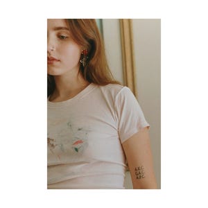 May include: A close-up shot of a person wearing a light pink t-shirt and a pair of earrings. The earrings are red and gold. The person has a tattoo on their arm that says "A.E.C. G.A.C. A.P.C."