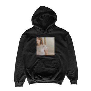 May include: Black hooded sweatshirt with a front pocket and a drawstring hood. The sweatshirt features a rectangular image on the front, depicting a person in a white t-shirt. The image has a soft, muted color palette.