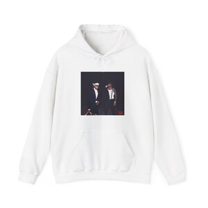 May include: White hooded sweatshirt with a front pocket. The image on the front of the sweatshirt features two people in suits. The background is black. The sweatshirt is made of a soft material.