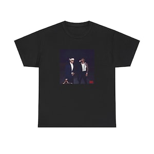 May include: Black t-shirt featuring a graphic of two people in suits and sunglasses. The image is set against a dark background. The t-shirt is made of a soft, comfortable material.
