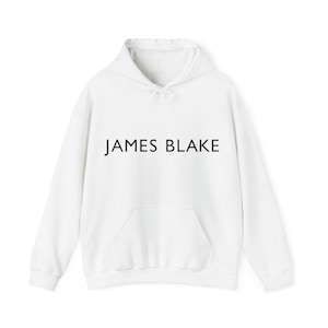May include: White hoodie featuring a front pocket and a drawstring hood. The words "JAMES BLAKE" are printed in black across the chest. A casual garment, suitable for everyday wear.