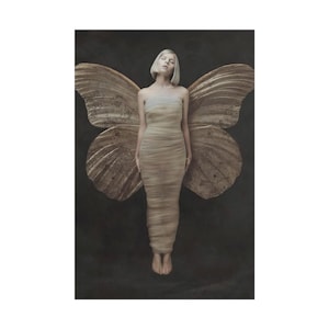 May include: A person with short blonde hair stands in front of large, textured butterfly wings. The person wears a beige, strapless dress. The wings and background are dark, creating a contrast with the person's light skin and hair.