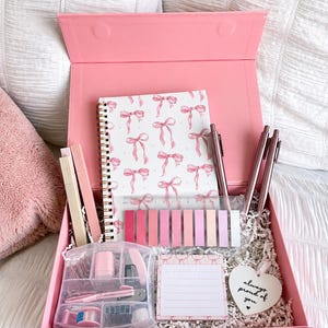 May include: A pink gift box containing a spiral-bound notebook with a pink bow pattern, three metallic pens, a clear ruler, a set of pink adhesive page markers, a clear organiser with stationery supplies, a lined notepad, and a heart-shaped tag.