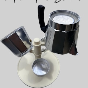 Peut inclure: Un support pour cafetière italienne de couleur crème avec une base circulaire et un support vertical central. Il maintient une cafetière Grosche argentée avec une poignée noire et un entonnoir séparé. Le texte Moka Pot Stand est visible.