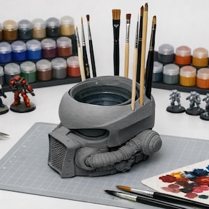 Pode incluir: Um suporte de pincéis cinzento em forma de capacete com um centro oco para guardar pincéis. O suporte apresenta uma grelha frontal texturada, tubos laterais e detalhes circulares. Assenta numa base de corte cinzenta com vários pincéis.