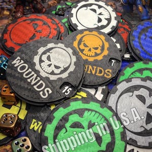 May include: A collection of circular plastic wound counters for tabletop gaming, featuring gear-shaped edges and skull icons in various colors including red, green, orange, and white. Each counter displays the text WOUNDS and a numerical value.