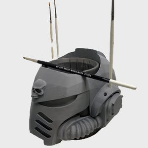 May include: A gray, 3D-printed helmet with a skull emblem and several paintbrushes. A black paintbrush with the text "DRY BRUSH MICRO-MARK 81081" is placed across the helmet. The helmet has a detailed, mechanical design.