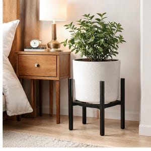 May include: A white textured ceramic planter with a green plant, set on a black metal plant stand. The stand has a four-legged design. The planter is near a wooden nightstand with a lamp and a clock.