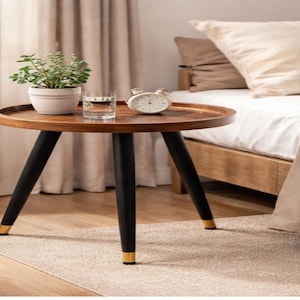 May include: A round wooden side table with black legs and gold accents. The tabletop holds a potted plant, a glass of water, and a small alarm clock. The table is near a bed with a beige comforter.